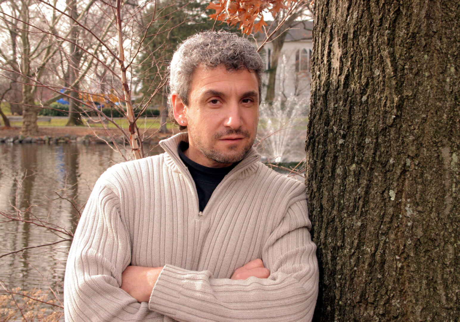 Man in a beige sweater leaning against a tree outdoors.