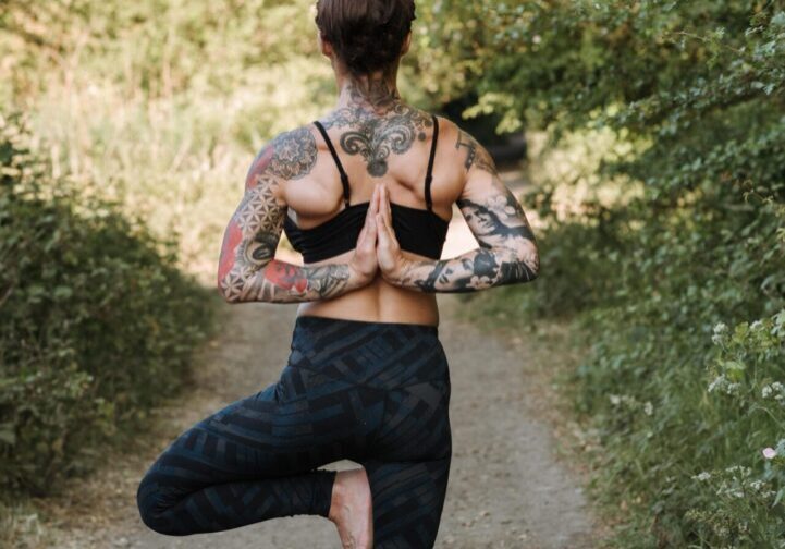 A tattooed woman practices yoga outdoors on a forest path.